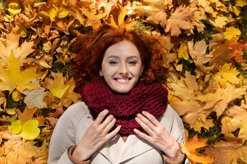 A person with red hair smiles while lying on a bed of vibrant autumn leaves. They wear a thick scarf and enjoy the warm colors of fall, creating a cheerful atmosphere.