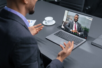 Two professionals are engaged in a video call discussing business matters. One person is showing gestures while speaking, and the other is smiling on the screen. A cup is placed nearby.