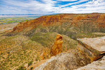 A narrow sandstone pinnacle rises from the canyon floor, surrounded by expansive layered cliffs in Colorado National Monument.