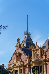 Intricate architecture of an old red brick building featuring a towering spire, ornate gables, and decorative sculptures under a vibrant blue sky.
