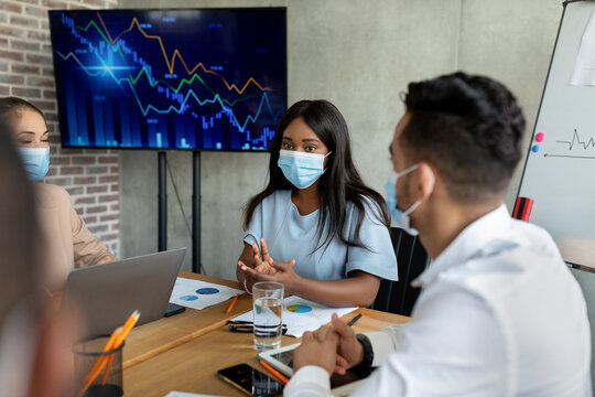 Multiethnic coworkers participate in a strategy meeting in a modern conference room. They wear protective face masks while discussing plans and analyzing charts on a screen. - Powered by Adobe