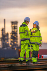 Two industrial workers in high-visibility safety uniforms discuss work plans on-site during golden hour, with a refinery or factory plant blurred in the background.