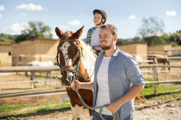 Happy father leads horse with his son on it - walk around a horse farm
