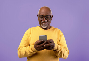 A man with gray hair and glasses holds a smartphone in his hands, focusing on the screen. He wears a yellow sweater and stands in front of a purple backdrop.