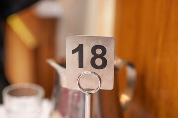 A detailed view of a restaurant table number '18' crafted from brushed metal, standing on a white tablecloth with soft, out-of-focus lighting.