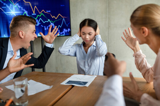 In an office conference meeting, several aggressive colleagues are shouting at a young Asian female employee. She appears upset and covers her ears, trying to protect herself from the verbal attack.