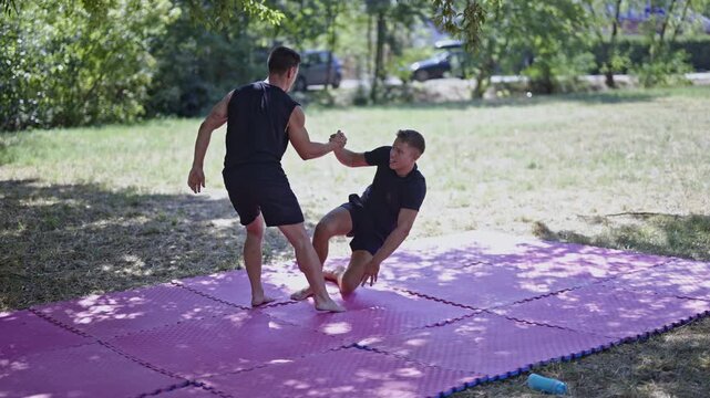 Two athletic men engage in intense wrestling and mixed martial arts combat training on a mat in an outdoor park. They practice takedowns and grappling, focusing on strength and physical fitness.