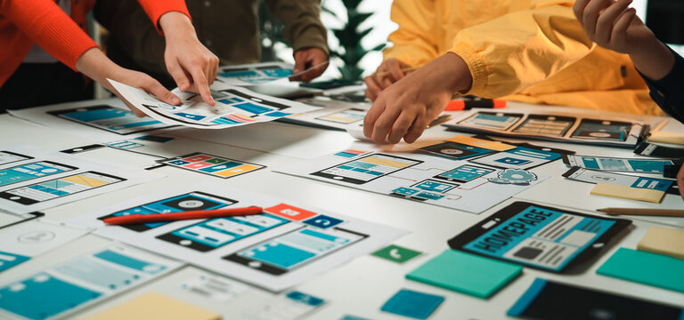 A group of individuals collaborates over a large table, analyzing wireframe mockups and colorful notes in a modern office setting, highlighting teamwork and creativity. SACTR