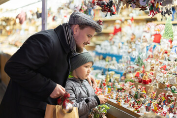 Dad and son are standing near counter at Christmas market fair, choosing figurines for nativity scene. Customers look at product with interest, select right figures. .