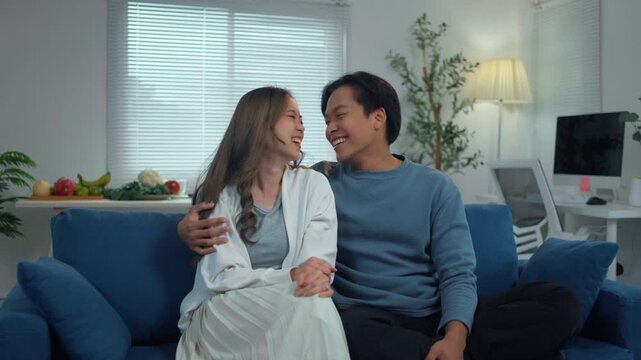 Happy young asian couple sitting on a blue sofa in their living room, talking, laughing, and showing affection with hugs and kisses