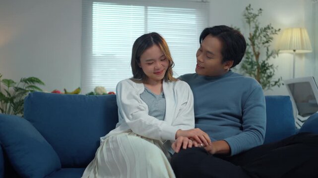 Happy young asian couple sitting on a sofa at home. Man and woman talking, laughing, and enjoying their time together in the living room