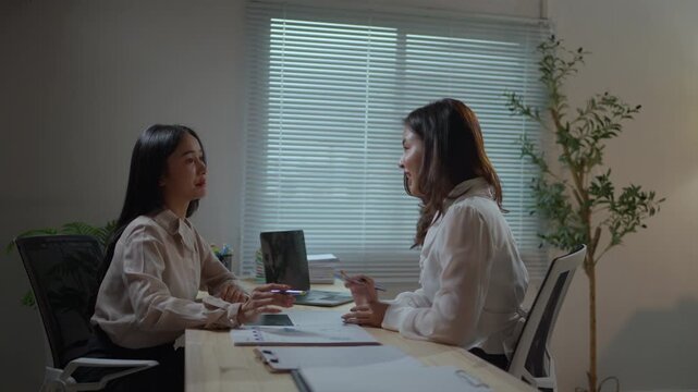 Two dedicated Asian businesswomen working late at night, collaborating on a project and analyzing documents together in a modern office