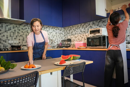 Two Asian women preparing healthy meal together in a modern blue kitchen, Sisters or friends organizing kitchen and setting the table with fresh fruits - Powered by Adobe