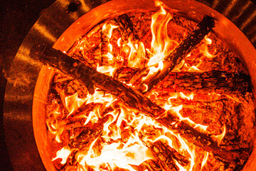 Intense flames engulf logs in an outdoor fire pit at night. The fire pit is circular with charred wood arranged in a cross pattern, surrounded by glowing embers warm orange and yellow hues and steel 