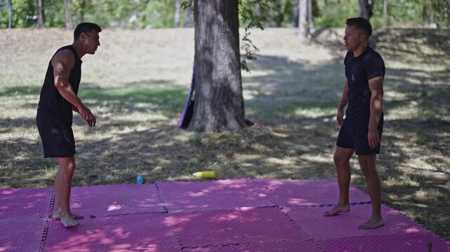 Two strong young men engage in intense mixed martial arts and wrestling training on pink mats in a sunny park. They practice combat techniques, focusing on grappling and physical fitness outdoors.