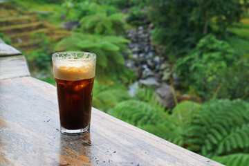 Iced americano placed on a wooden table on the edge of a cliff with a view of the green valley in the distance