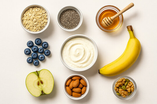 Flat lay of wholesome breakfast ingredients including yogurt, blueberries, banana, apple, almonds, walnuts, chia seeds, oats, and honey arranged neatly on a clean white background