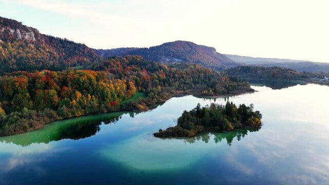 Slow aerial pull out over Lac d&rsquo;Ilay and small wooded islet with turquoise shallows along shore and hills of Jura Mountains near Le Frasnois and La Chaux‑du‑Dombief, Bourgogne‑Franche‑Comt&eacute;, France