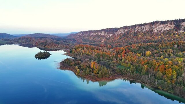 Lac d&rsquo;Ilay reflecting sky, small forested islet and a curving shoreline of autumn woodland, limestone Jura cliffs and plateau near La Chaux‑du‑Dombief in Haut‑Jura, Bourgogne‑Franche‑Comt&eacute;, France
