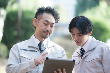 新緑の屋外の施設管理など現場で働く作業着を着た施工管理士などのビジネスマン　タブレットを見る