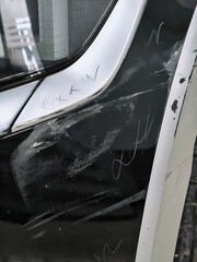 A vertical close-up of a damaged car's bumper panel showing deep scratches, scuffs, and assessment markings in marker used by a repair shop or insurance adjuster to prepare an estimate and claim