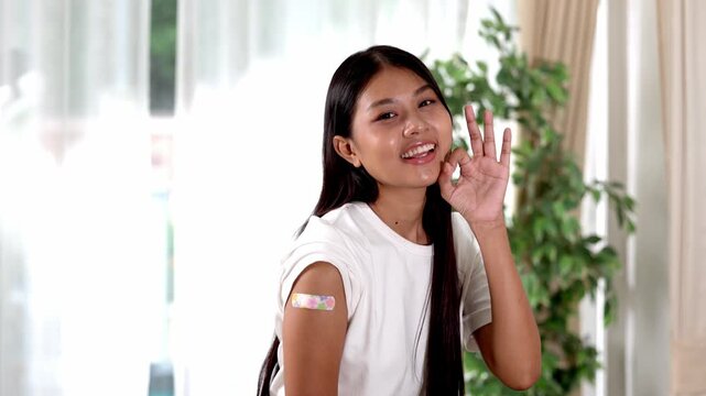 Asian Young woman with bandage upper arm after vaccination smiling and making OK gesture, adhesive vaccine patch visible as she stands indoors bright room expressing calm confident immunity