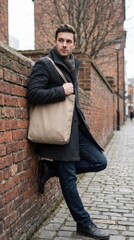 Obraz premium Stylish man in black coat and jeans leaning against a brick wall with a tote bag.