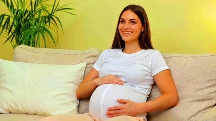 Pregnant woman smiling while holding her belly sitting on a couch indoors happy maternity lifestyle expecting mother health care prenatal joy relaxation domestic scene beautiful expectant parent comfo