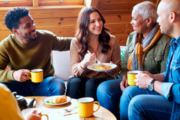 Group of young adult and middle aged multiethnic men and women sitting together, smiling and talking while eating breakfast and drinking coffee in cozy wooden room, enjoying casual conversation