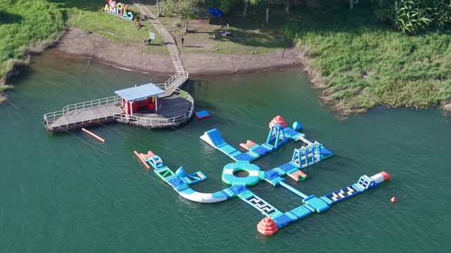 Drone video captures a vibrant floating water park on Lago Calima. Set in peaceful green surroundings, the colorful inflatable structures create a lively atmosphere on the serene lake waters.