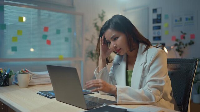 Young Asian woman feeling stressed and frustrated while working late at her desk. Overworked professional suffering from a headache