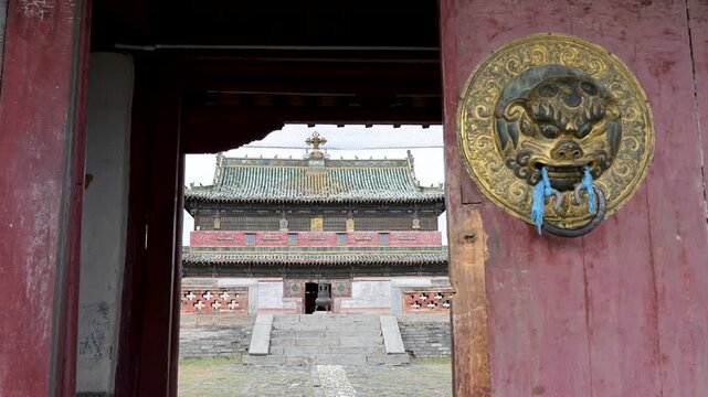 Looking through a historic gateway at Erdene Zuu. The main Buddhist temple with its green tiled roof is revealed behind a traditional brass door handle and ceremonial scarf.