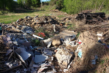 Outdoor illegal dump site featuring construction debris, broken tiles, soil, and dry brush in a green, overgrown area in spring