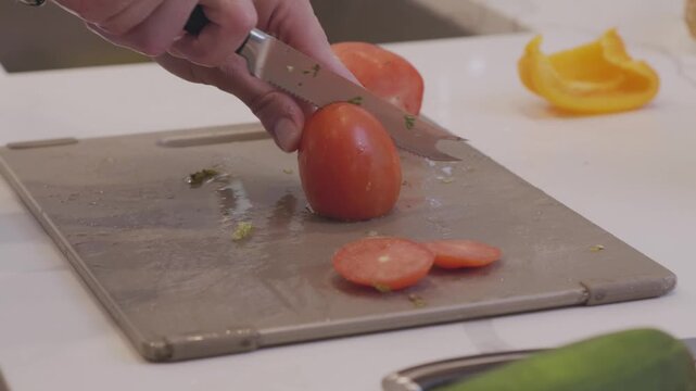 A close up of slicing roma tomatoes on a cutting board surrounded by vegetables in a modern kitchen.