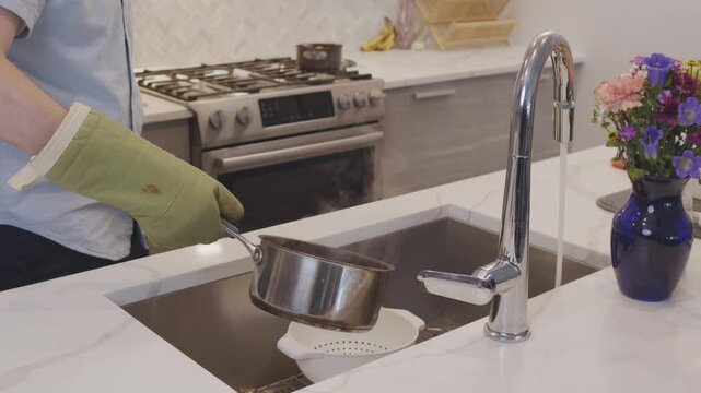 A slow motion shot of a pot of steaming hot pasta being poured into a strainer in a home kitchen sink.