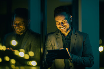 Man in formal attire smiling while using a tablet in a softly lit modern setting at night