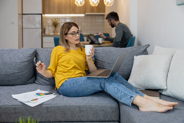 Young woman working on laptop while sitting on a couch with a cup in a cozy home office setting