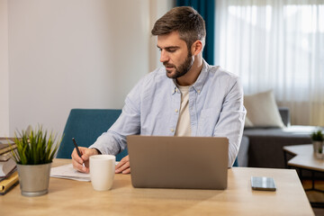 Man working on laptop while writing notes at home in a bright and modern living space