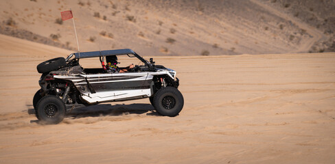 White and black dune buggy racing across sunlit sand dunes, kicking up dust in a high-speed off-road desert adventure under clear skies. © Daniel Wedeking