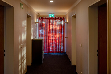 Hallway of a hotel with an emergency exit sign and the morning sun shining through a red semi translucent curtain. 