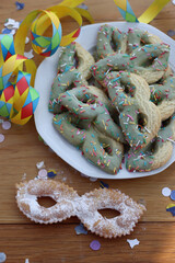 Many cookies in the shape of a Carnival mask with Pistachio glaze ith confetti and paper serpentine and Carnival mask hape crostoli on table