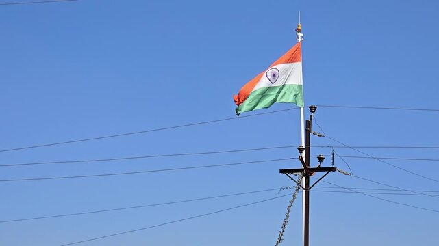 A low-angle view of the Indian flag flying high against a clear blue sky. The tricolor flag, with saffron, white, and green bands, and the central Ashoka Chakra, above utility lines or electric pole.