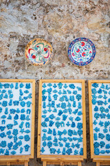 Beautiful display of traditional Turkish blue ceramic amulets and handcrafted decorative tiles arranged in wooden frames against a rustic stone wall. Featuring iconic evil eye symbols, floral motifs