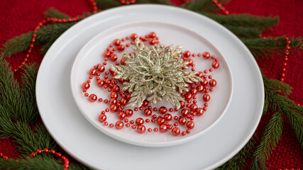Festive holiday table setting featuring a white plate adorned with a golden floral centerpiece and red beads, surrounded by evergreen branches, creating a warm seasonal atmosphere