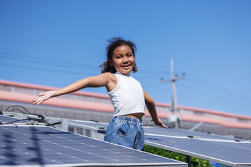 A joyful young girl stands with open arms beside solar panels under a bright blue sky, expressing...