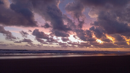 A breathtaking sunset over a peaceful beach featuring dramatic purple clouds, rolling ocean waves, and a soft golden horizon glow