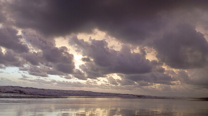 ​A moody coastal scene featuring heavy dark storm clouds over a reflective beach, with crashing white waves under dim light