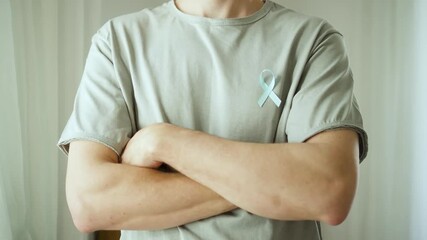 A young man with a blue satin ribbon on his tshirt stands with arms crossed. November is Prostate Cancer Awareness Month for diagnosis and support.