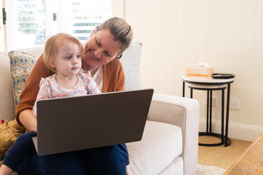 Mother working on her laptop with toddler daughter sitting on her lap on the couch