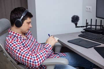 Man wearing headphones sitting at an ergonomic desk, writing notes in a notebook